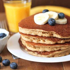 Four banana pancakes on a white plate with fresh slices of banana and blueberries on top.