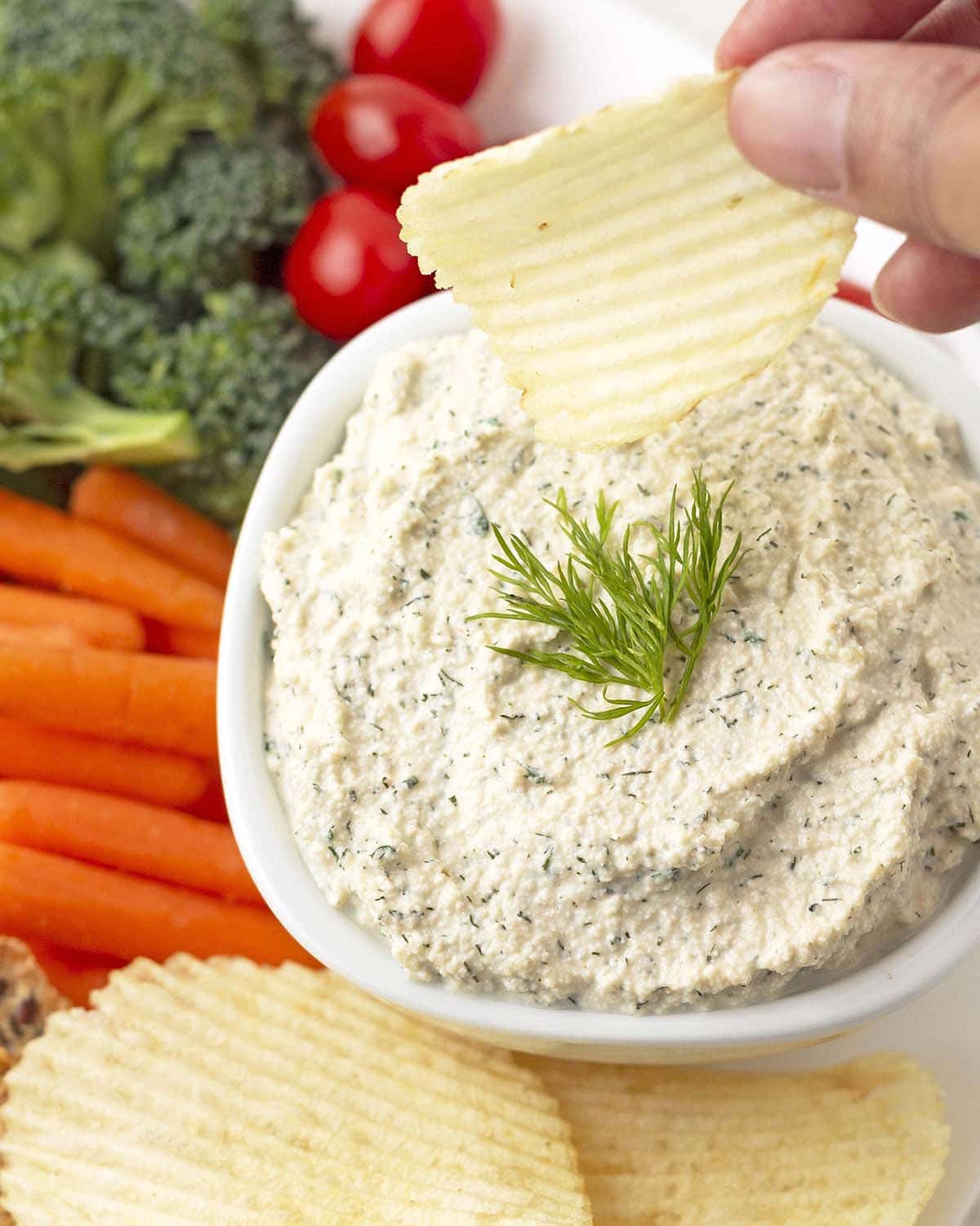 A hand dipping a potato chip into a bowl of dip, veggies and chips surround the bowl.