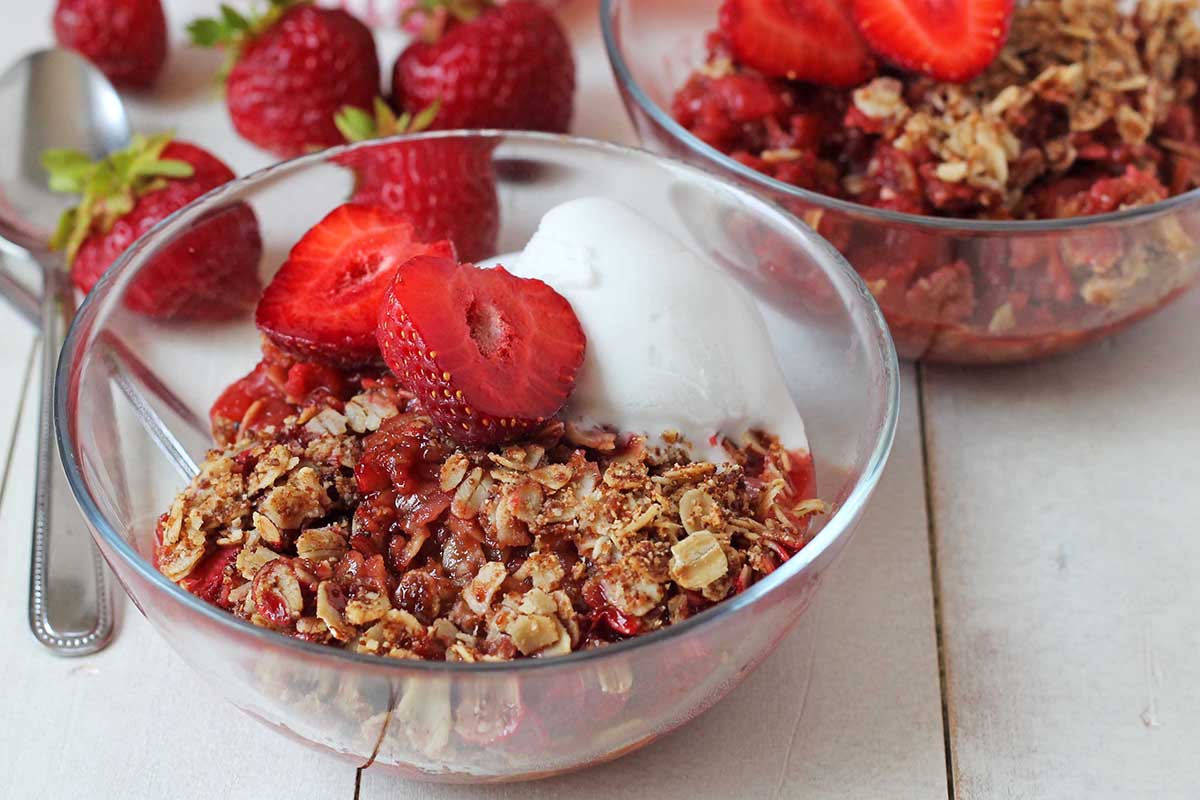 A small glass bowl filled with strawberry rhubarb crisp, it is topped with a halved strawberry and coconut ice cream.