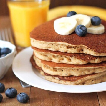 A stack of four banana pancakes sitting on a white plate, pancakes are topped with sliced bananas and fresh blueberries.