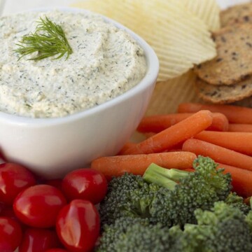 A bowl of dill dip on a plate, the plate is filled with fresh vegetables, chips, crackers, and pretzels for dipping.
