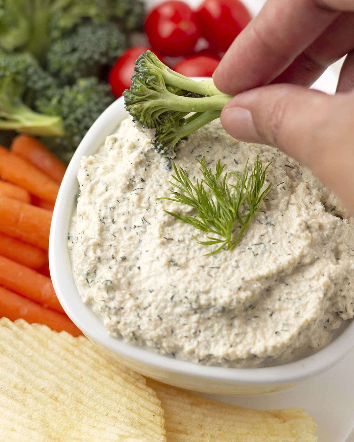 A hand dipping a piece of fresh broccoli into a bowl of dip.