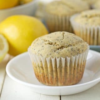 A muffin sitting on a white plate, fresh lemons sit behind the plate as well as another plate of muffins.