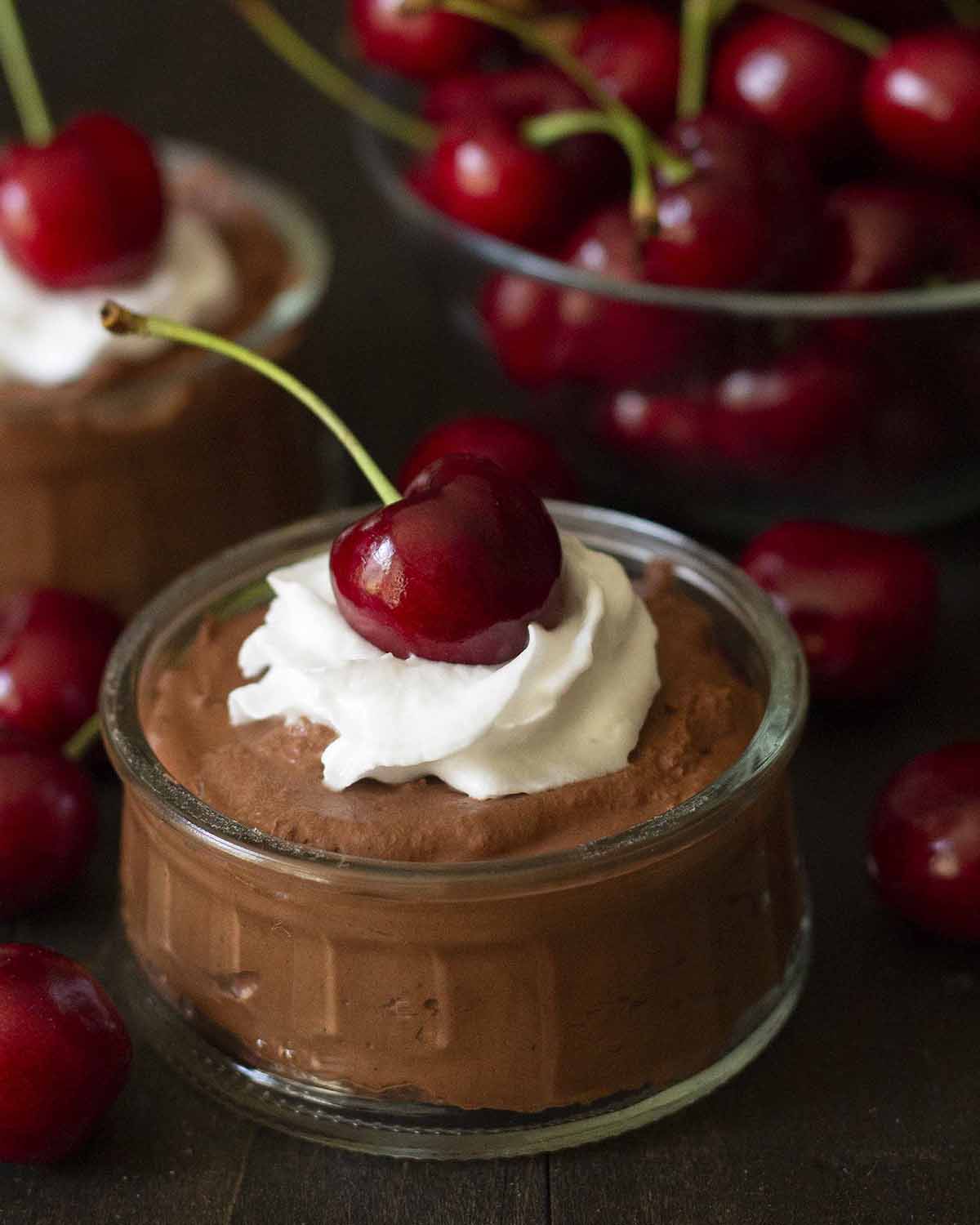 A close up shot of vegan chocolate mousse in a glass bowl, mousse has whipped cream and a cherry on top.