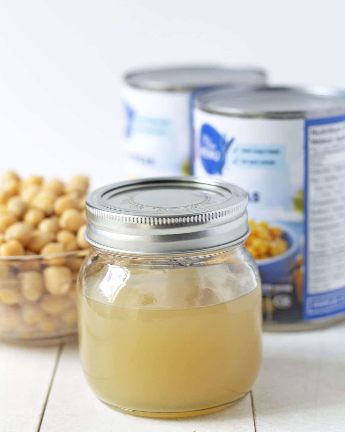 A jar of aquafaba egg replacement sitting on a white table, two chickpea cans and a bowl of chickpeas sit behind the jar.