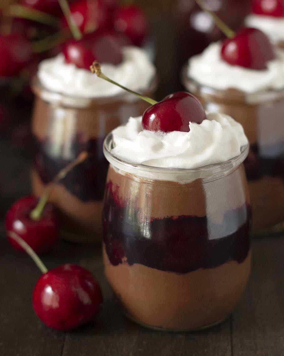 A dessert glass filled with vegan chocolate mousse layered with cherry sauce and whipped cream, and topped with a cherry.