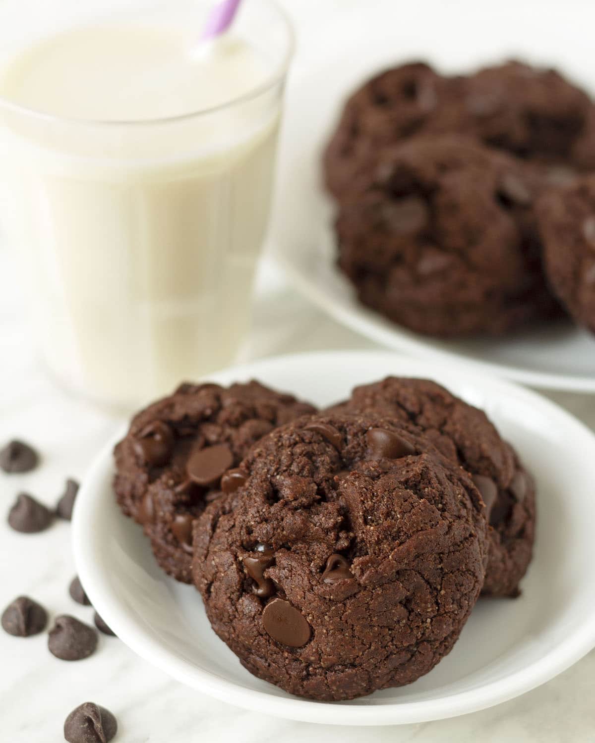 Three chocolate cookies on a small plate, a plate full of cookies and a glass of almond milk sits behind the plate.