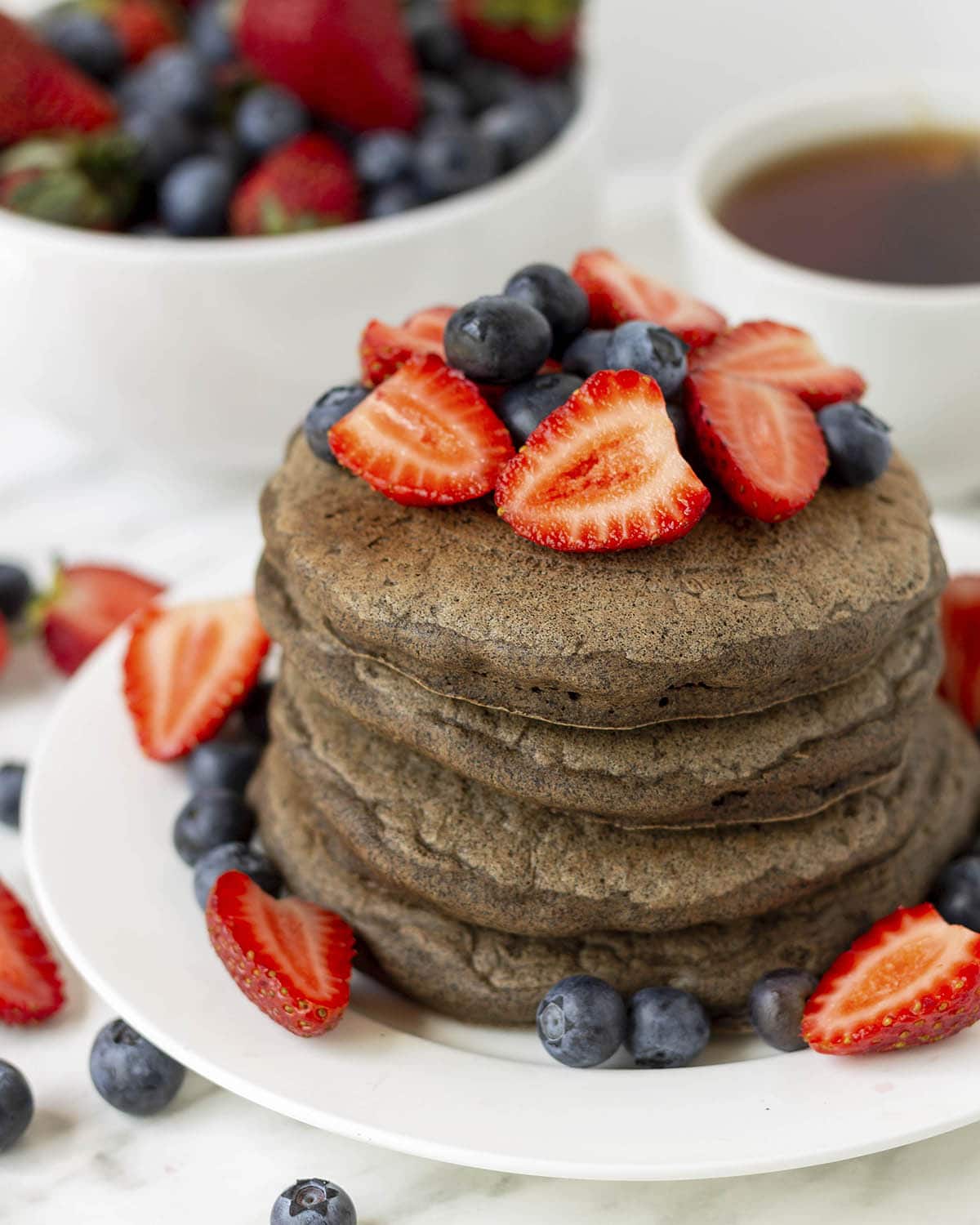 A stack of four buckwheat pancakes on a white plate, fresh blueberries and strawberries garnish the pancakes and plate.