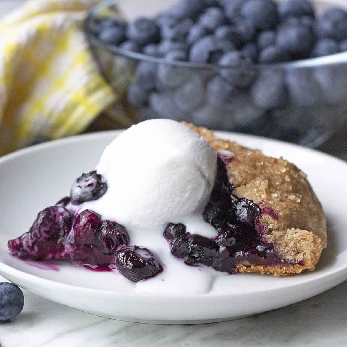 A slice of blueberry galette on a plate with a melting ice cream on top of it, a bowl of berries sits behind the plate. 