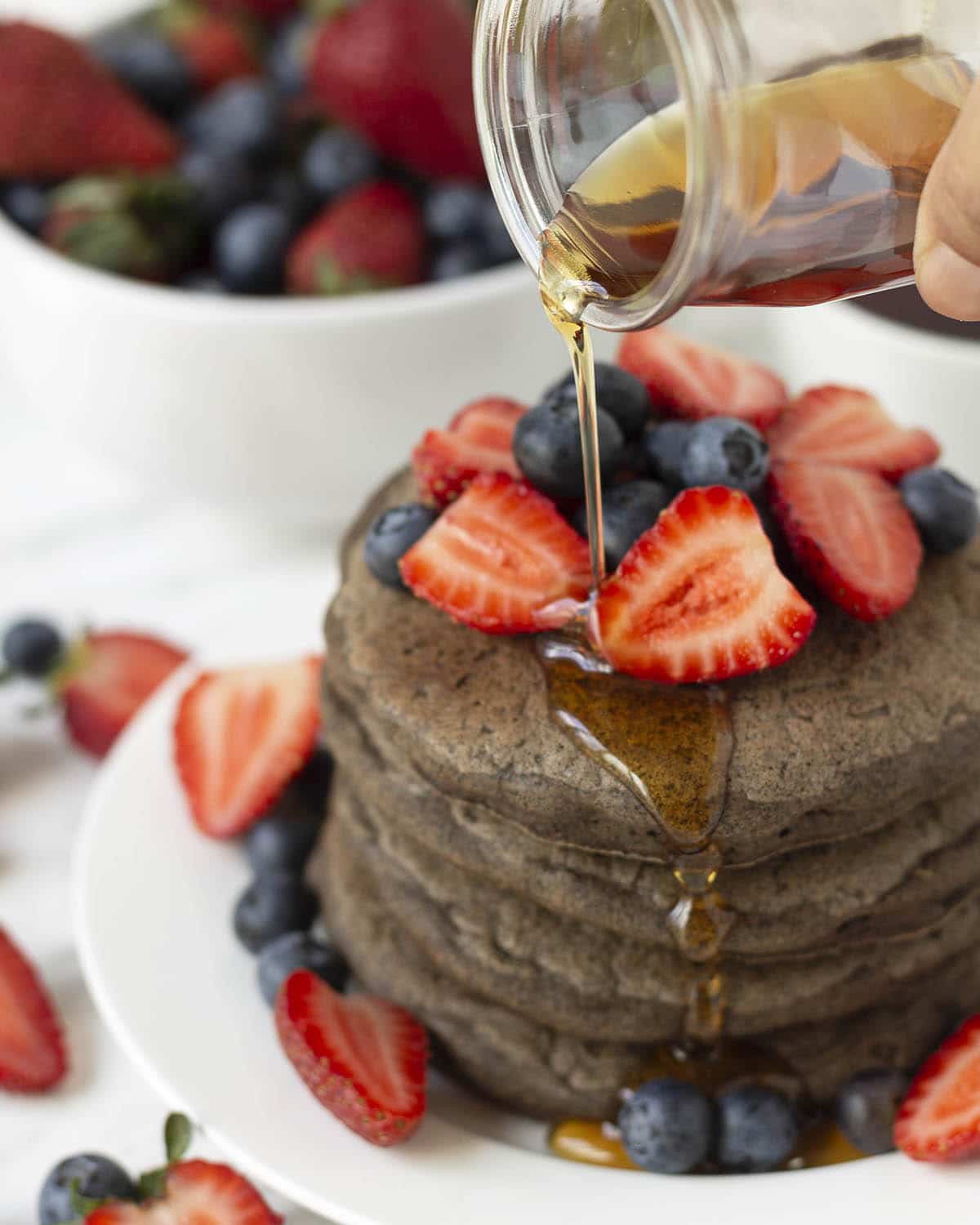 Maple syrup being poured from a small glass jar onto a stack of pancakes that are garnished with fresh berries.