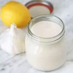 A small opened jar of vegan garlic aioli, a bulb of fresh garlic and a fresh lemon sits behind the jar.