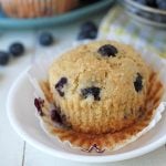 A blueberry muffin on a white plate, muffin wrapper is peeled down.