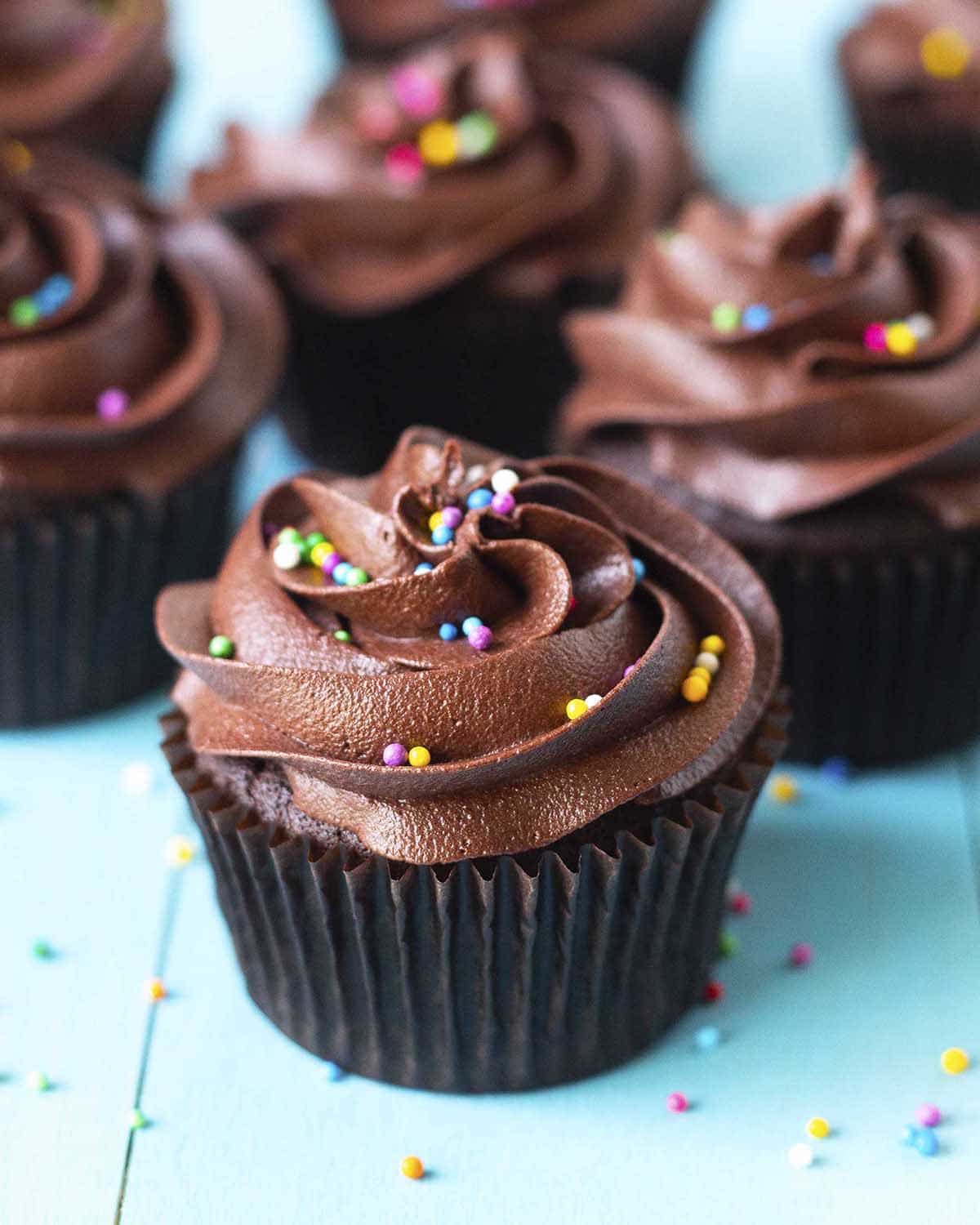 Several frosted chocolate cupcakes with sprinkles on top sitting on a blue table.