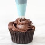 A blue piping bag and frosting tip piping chocolate buttercream onto a chocolate cupcake.