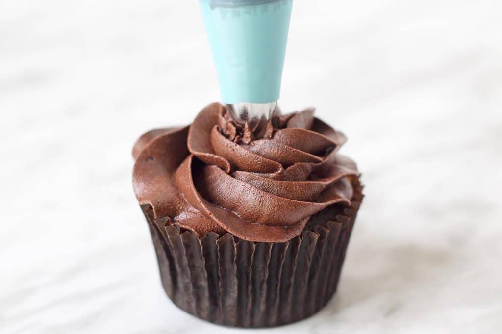 A frosting piping bag piping chocolate buttercream onto a chocolate cupcake.
