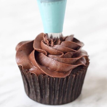 A frosting piping bag piping chocolate buttercream onto a chocolate cupcake.