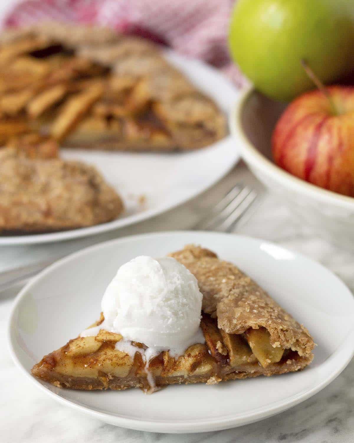 A slice of apple galette on a plate with a scoop of ice cream on top of it, a bowl of fresh apples sits behind the plate.