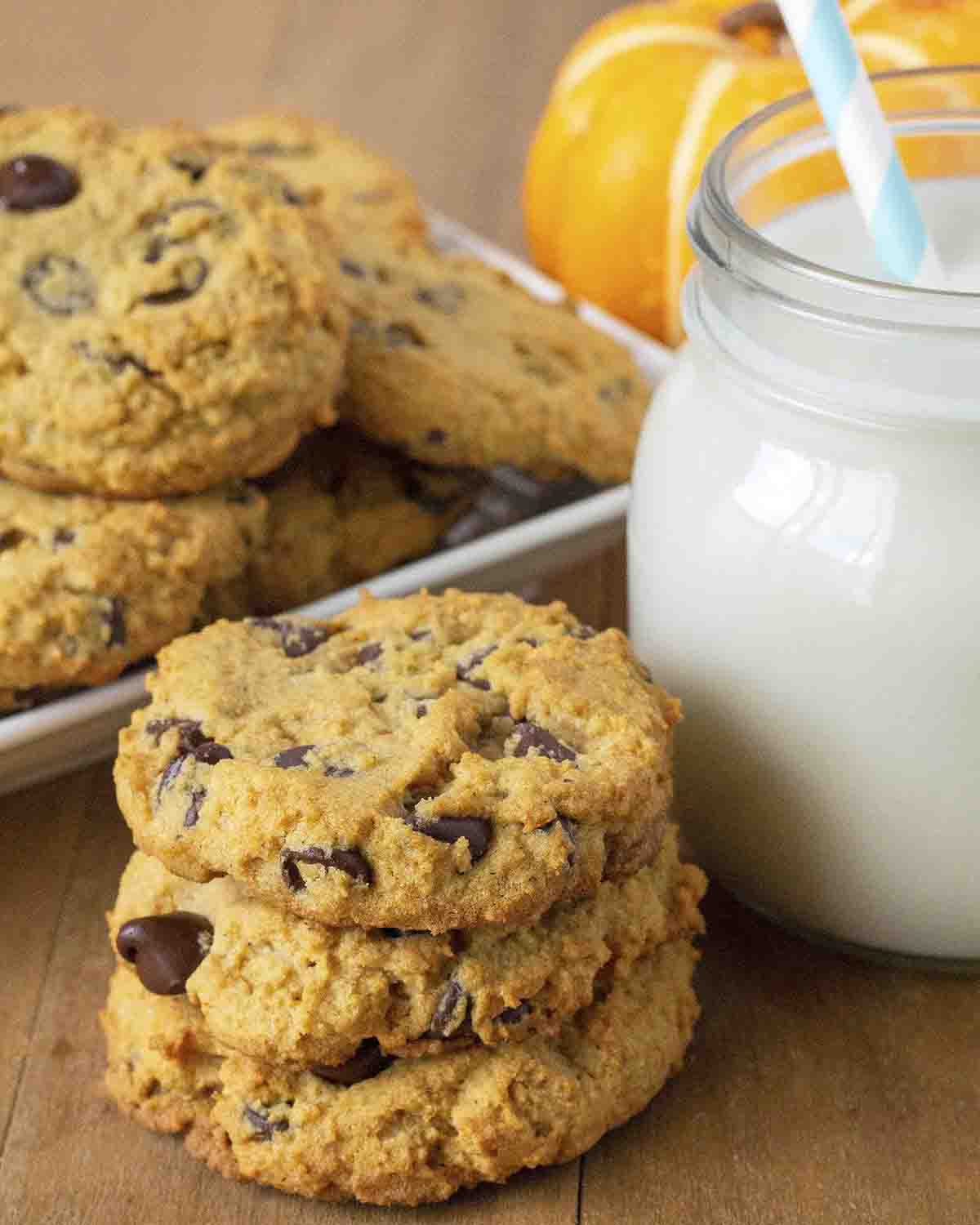 A stack of three vegan gluten free pumpkin chocolate chip cookies on a wood table, a mason jar of milk sits to the right.