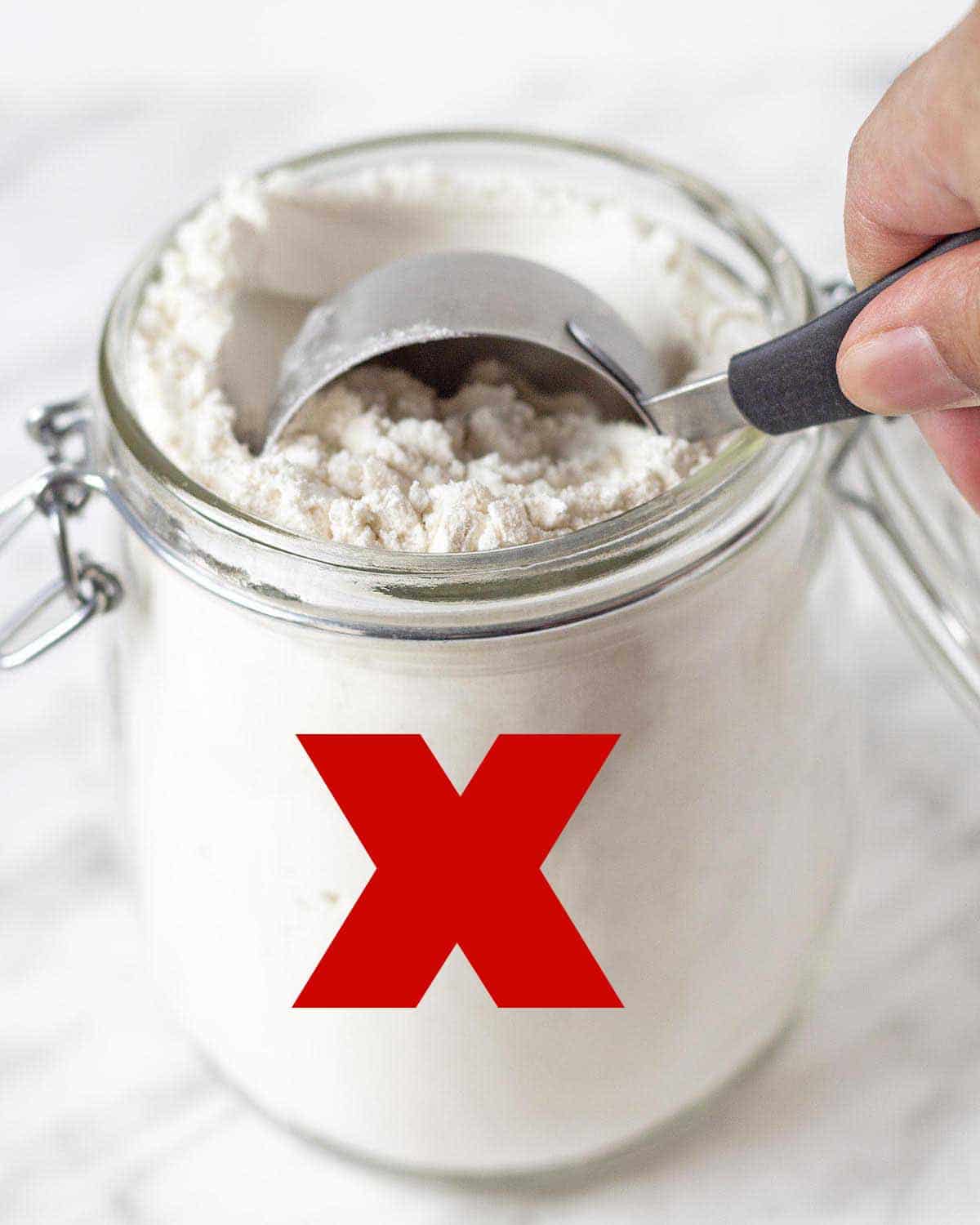 A hand scooping flour from a jar with a measuring cup, a red x is on the jar to show this is the wrong way to measure flour.