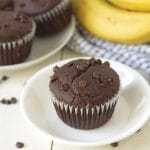 A chocolate banana muffin on a small white plate, fresh bananas and more muffins are in the background.