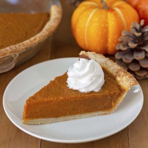 A slice of pumpkin pie on a white plate, pie has whipped cream on top, small pumpkins and pinecones are in the background.