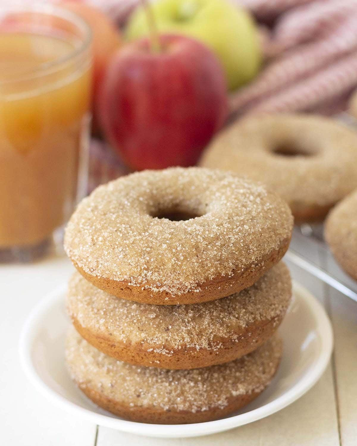 Three dairy free apple cider donuts stacked on top of each other, fresh apples and more donuts sit in the background.