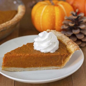 A slice of pumpkin pie on a white plate, small pumpkins, pinecones, and the rest of the pie are in the background.