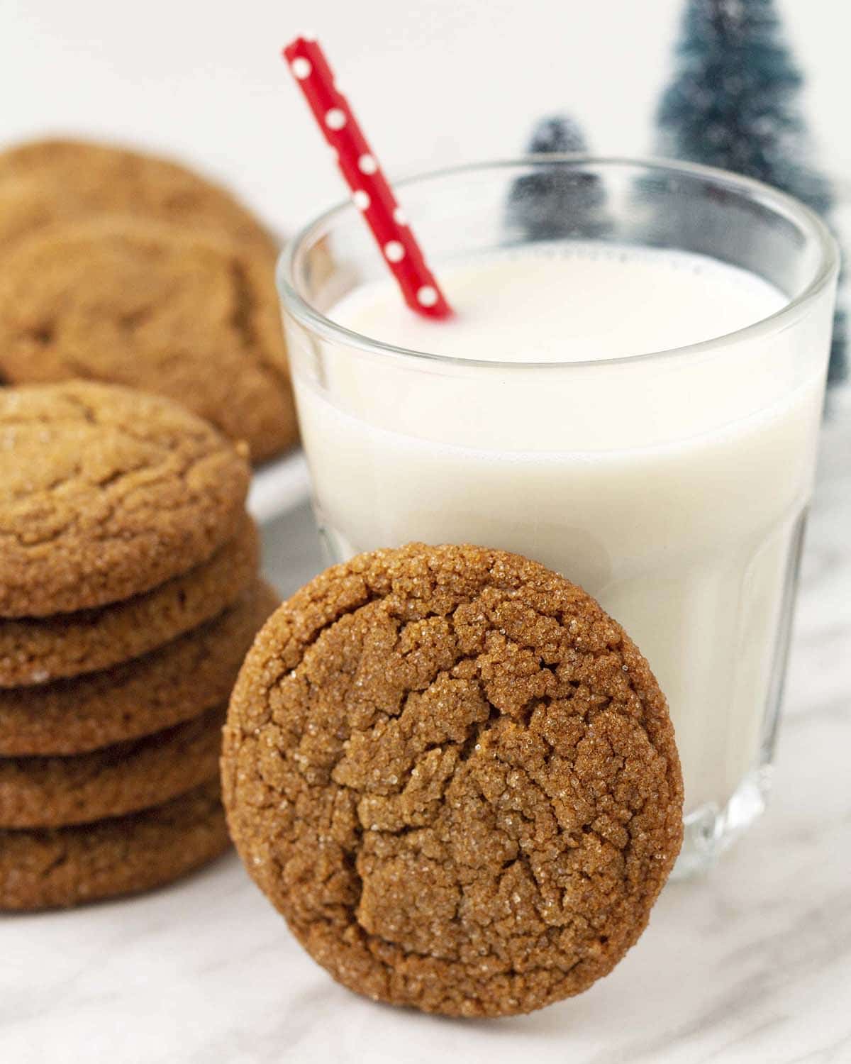 A glass of milk with a red straw, a molasses ginger cookies is leaning on it, more cookies sit in the background.