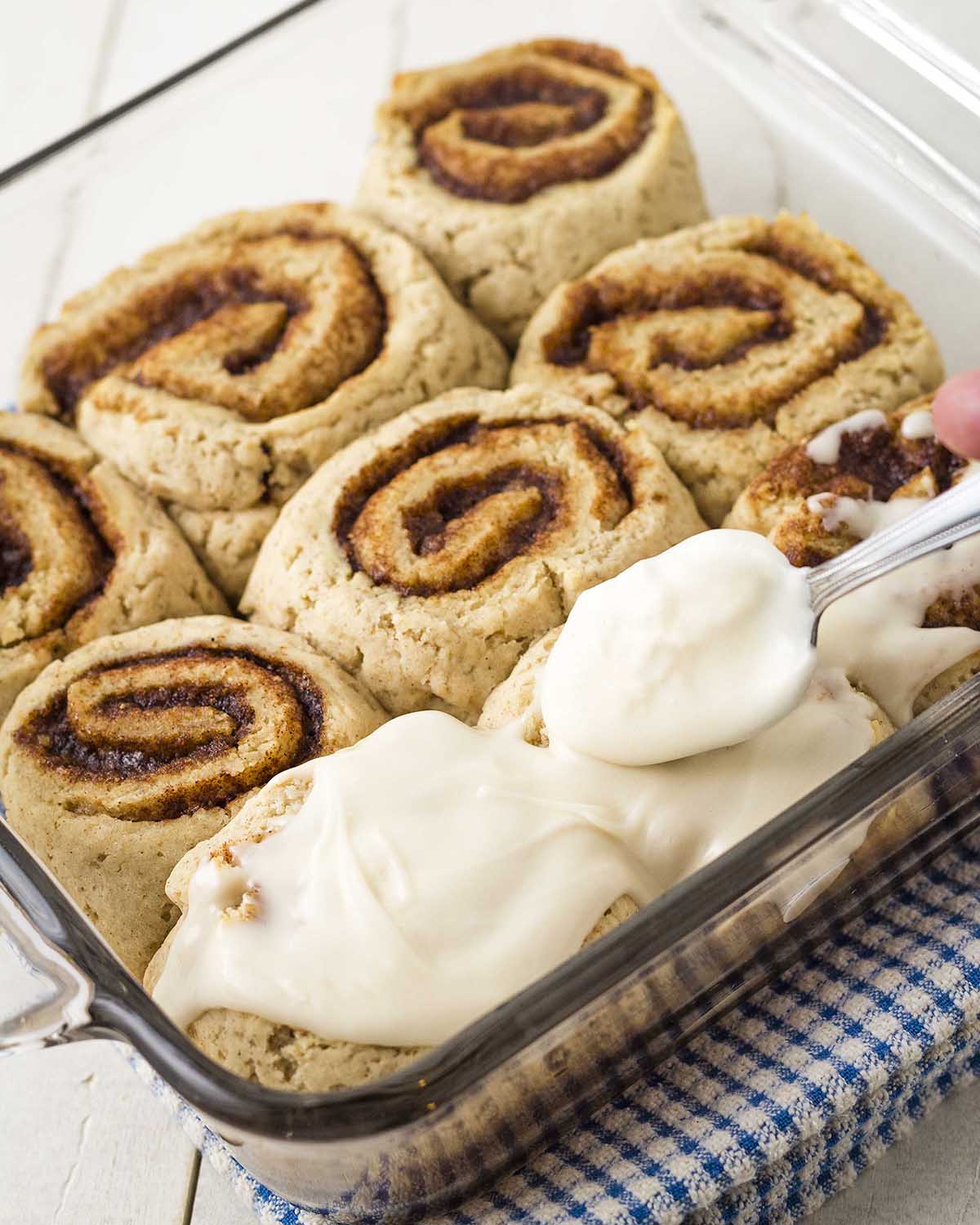 A baking dish of vegan gluten free cinnamon buns in a dish, icing is being added to the top of the buns with a spoon.