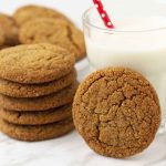 A stack of vegan molasses cookies on a table, one cookie is leaning against a glass of milk, a plate of cookies sits behind.