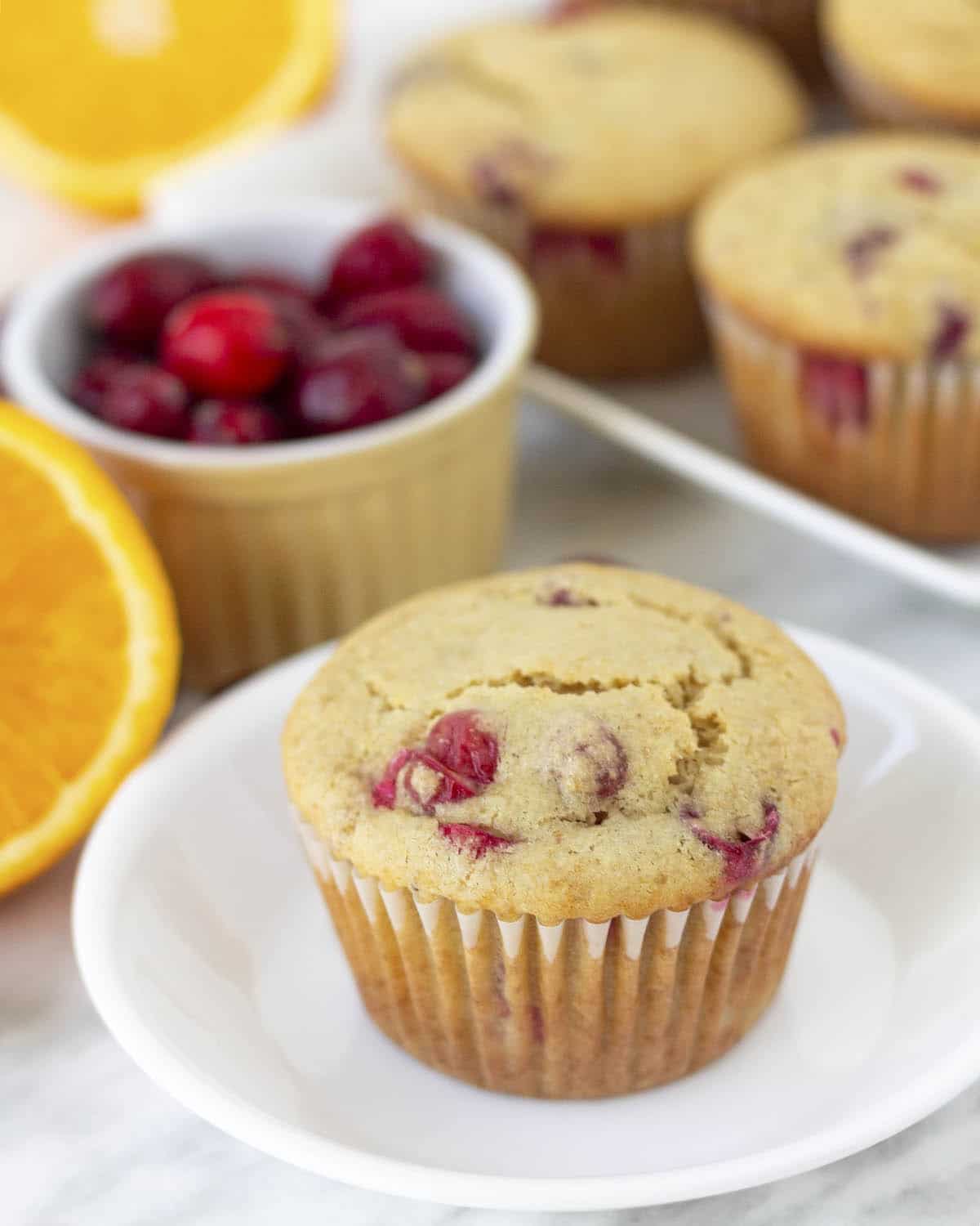 A cranberry orange muffin on a white plate, more muffins sit in the background as well as an orange and fresh cranberries.