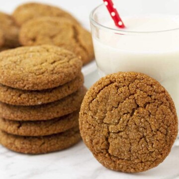 Egg free molasses cookies stacked on a table, one is leaning against a glass of almond milk, a plate of cookies sits behind.
