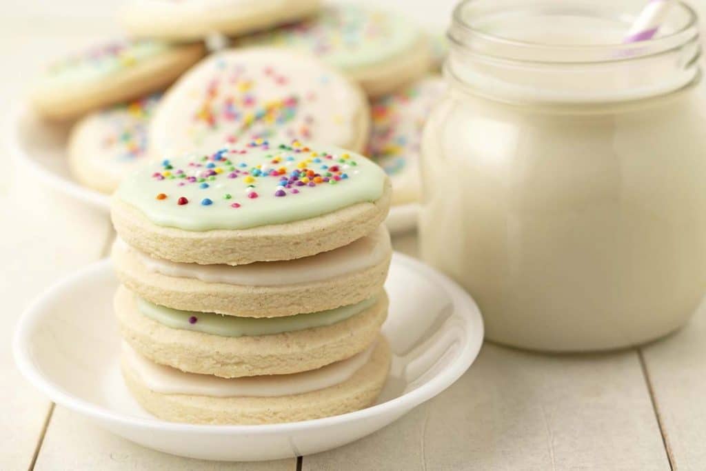A stack of four gluten-free sugar cookies on a small white plate, more cookies and a glass of milk sits behind it.