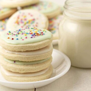 A stack of four gluten-free sugar cookies on a small white plate, more cookies and a glass of milk sits behind it.