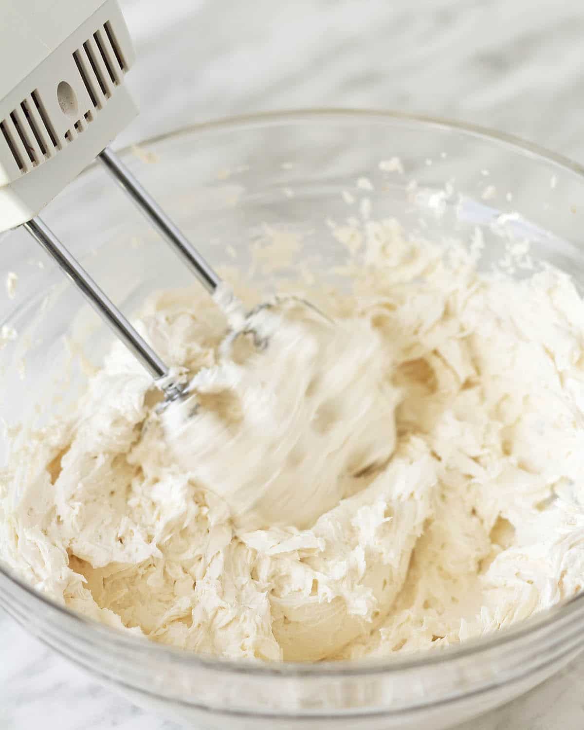 Image shows an electric beater whipping vanilla frosting in a glass bowl.