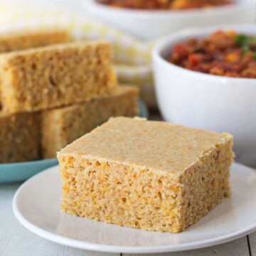 A square piece of cornbread on a white plate, two bowls of chilli sit in the background as well as a plate of more cornbread.