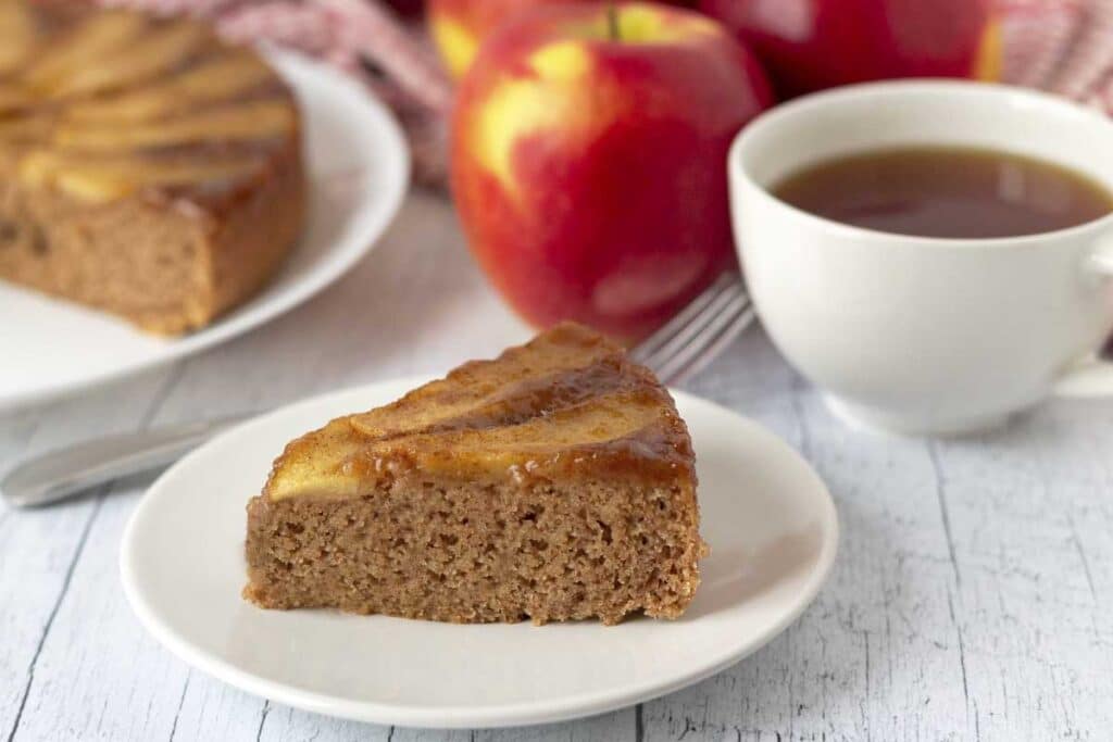 A piece of gluten-free apple upside down cake on a small white plate, fresh apples sit behind the plate.