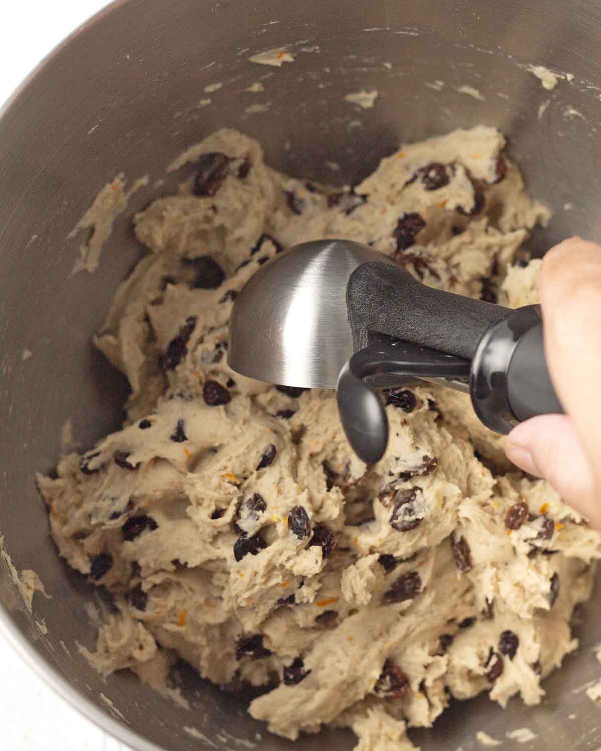 A hand scooping gluten-free bun dough with an ice cream scoop from a mixing bowl to form into rolls.