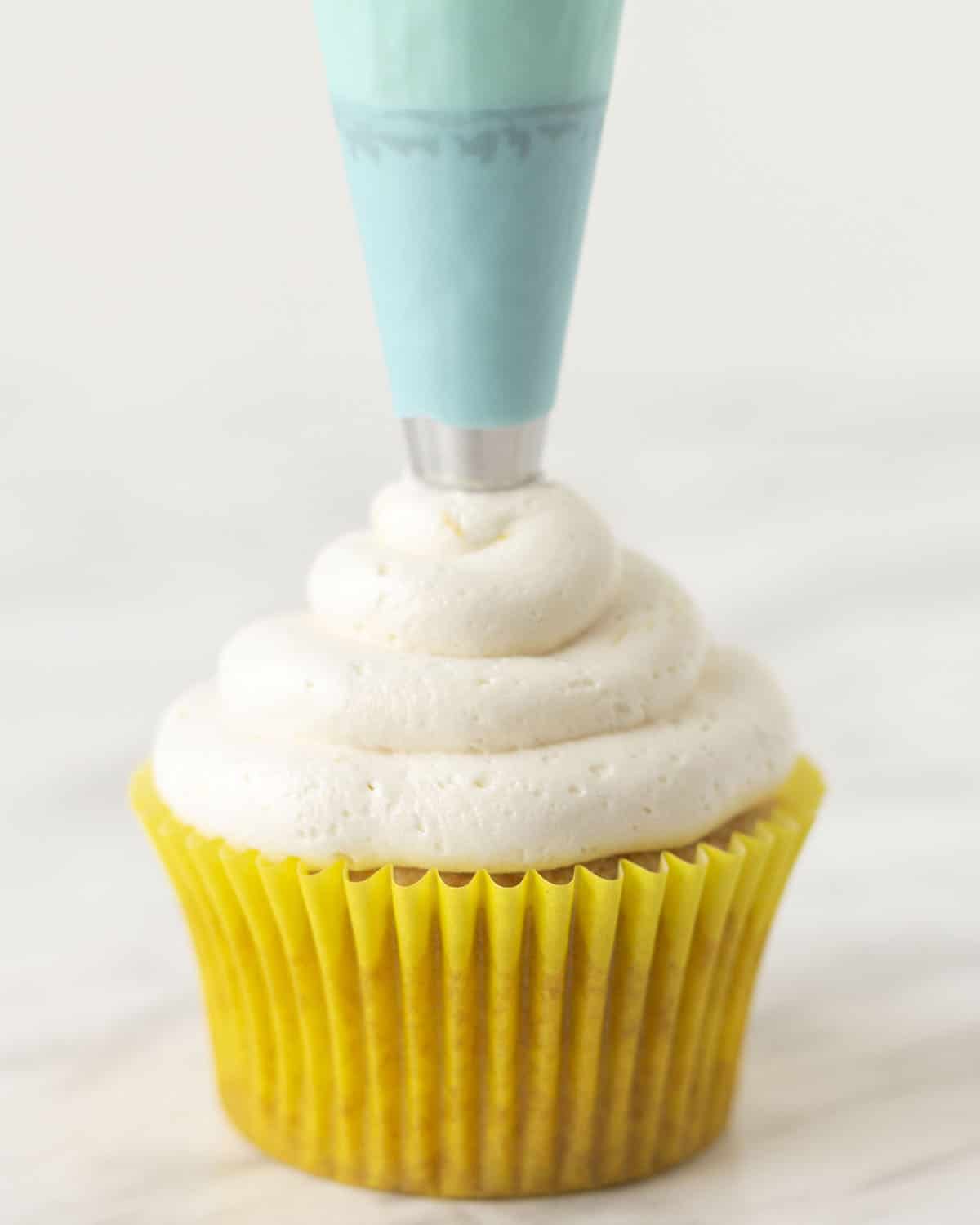 Vegan lemon buttercream frosting being piped onto a lemon cupcake with a blue piping bag.