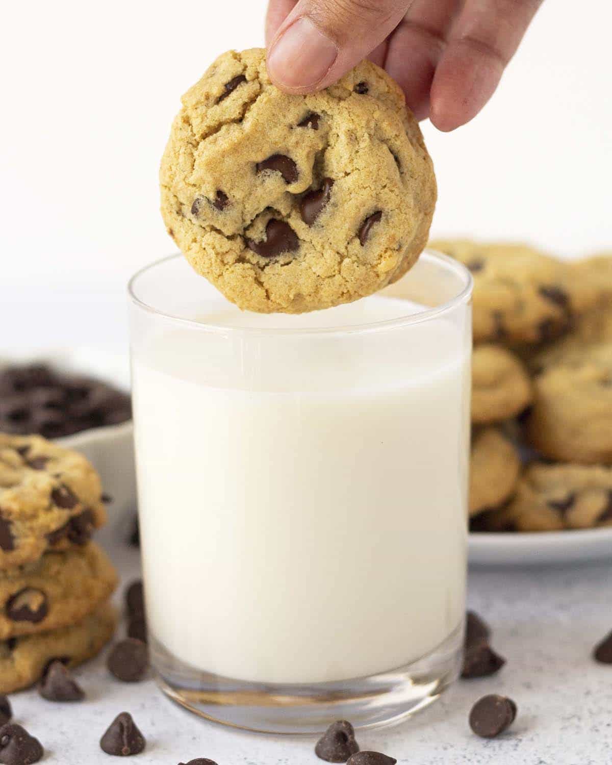 A hand holding a gluten-free chocolate chip cookie above a glass of almond milk.