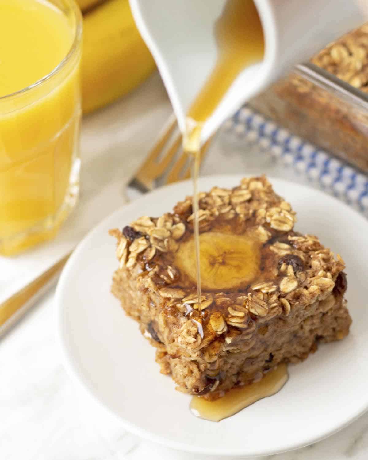 A slice of vegan banana baked oatmeal on a white plate, syrup is being poured onto the oatmeal.