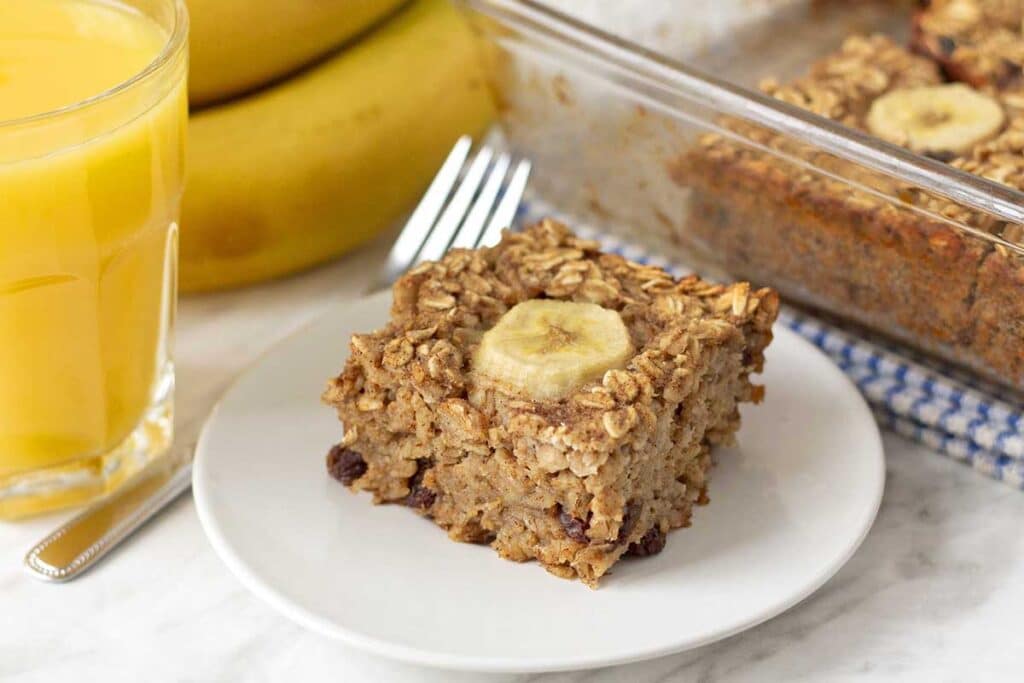 A piece of baked banana oatmeal on a white plate, a fork, a glass of orange juice, and bananas sit to the left of the plate.