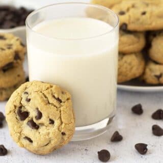 A chocolate chip cookie leaning against a glass of almond milk, more cookies sit on a plate behind the almond milk.