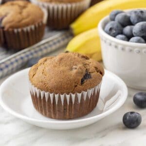 A muffin on a small white plate, fresh bananas, blueberries and more muffins sit in the background.