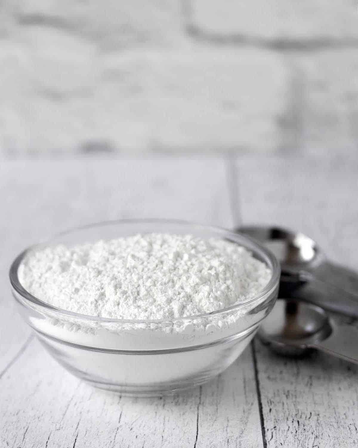 A glass bowl filled with baking powder, several metal measuring spoons sit to the right of the bowl.