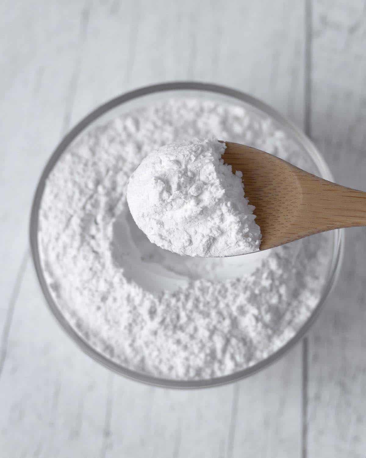 A wood spoon holding a spoon full of baking powder over a glass bowl filled with baking powder.