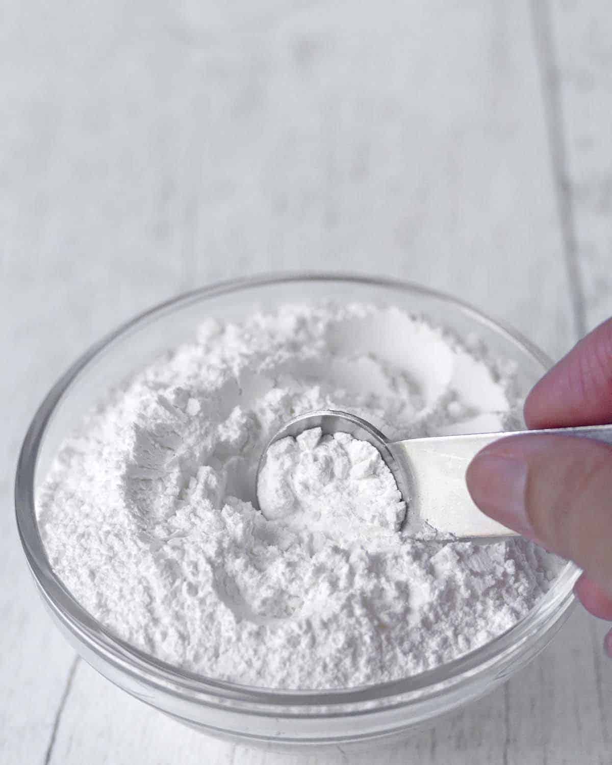 A hand scooping out baking powder from a glass bowl using a measuring spoon.