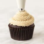 Peanut frosting being piped onto a chocolate cupcake with a cream coloured piping bag.