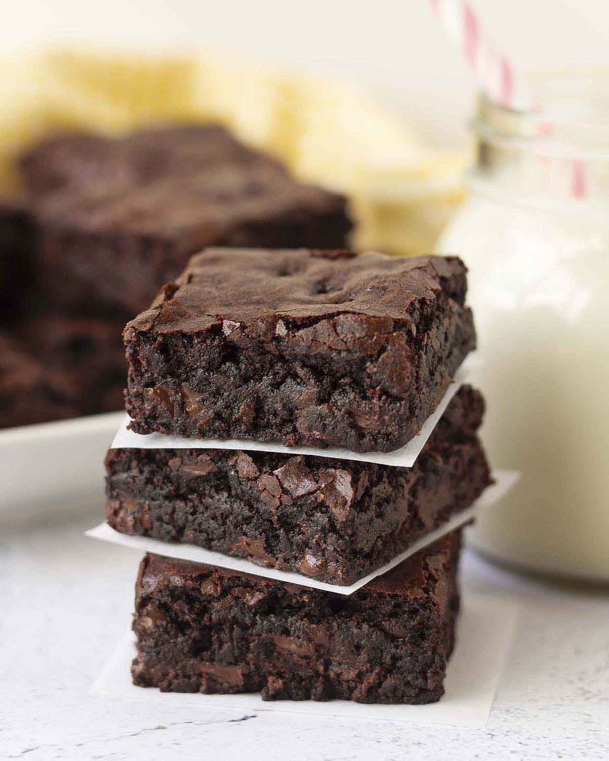 A stack of three gluten-free brownies, a glass of milk with a pink straw in it and more brownies sits behind the stack.