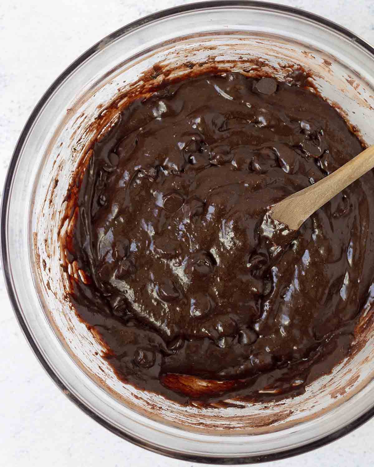 An overhead shot of a glass bowl filled with eggless brownie batter, there is a wooden spoon in the bowl.